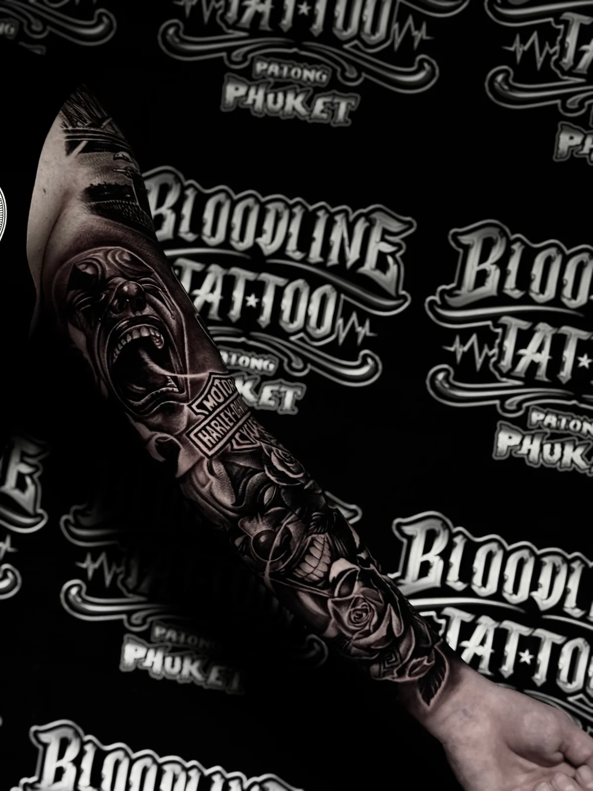 Chicano arm sleeve with screaming portrait, Harley-Davidson logo, payasa face, and roses at Bloodline Tattoo Phuket.