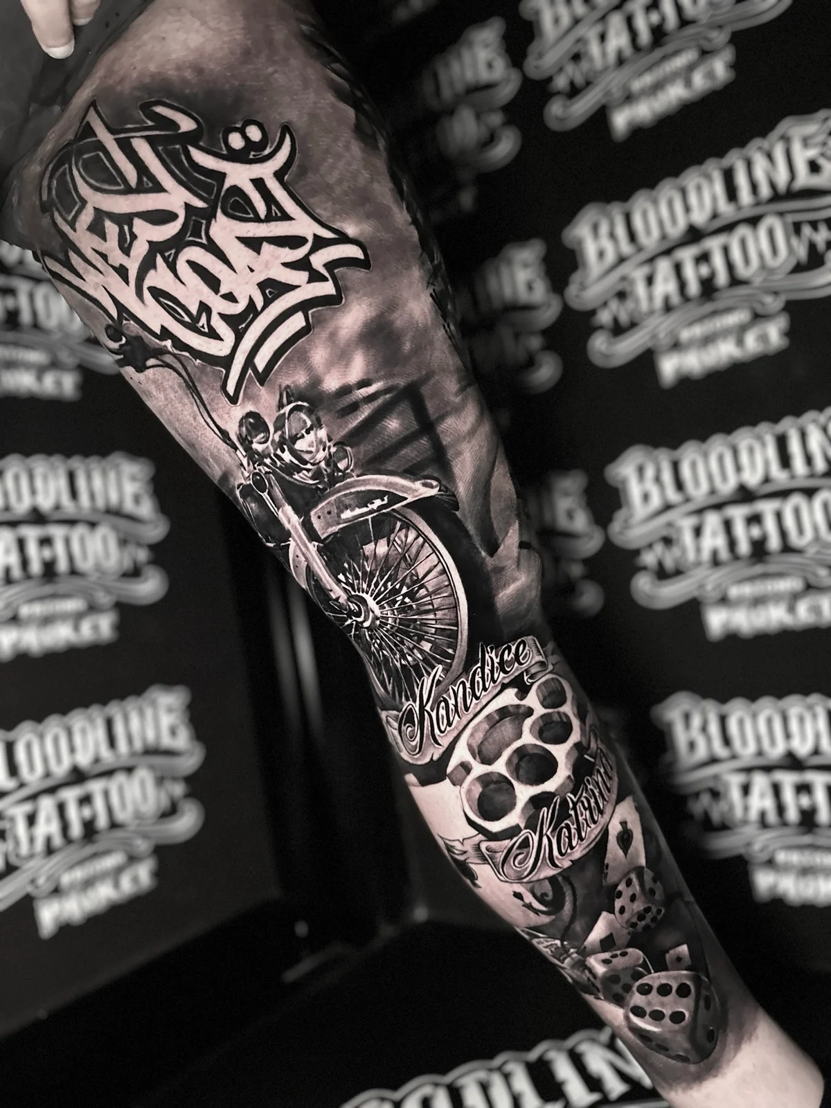 Chicano West Coast script leg tattoo with motorcycle, name banners, brass knuckles, and dice at Bloodline Tattoo Phuket.