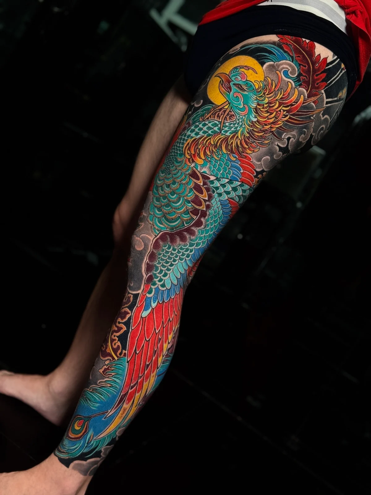 Japanese colour phoenix full-leg tattoo with teal scales, red and yellow tail feathers, and dark cloud backgrounds at Bloodline Tattoo Phuket.