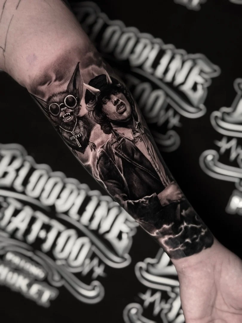 Black and grey portrait realism forearm tattoo of Angus Young with horned devil styling beside an Ozzy bat with lightning at Bloodline Tattoo Phuket.