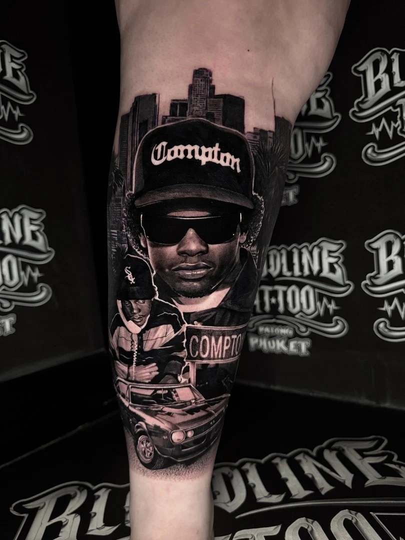 Black and grey portrait realism leg tattoo of Eazy-E with Compton street sign, skyline, and lowrider scene at Bloodline Tattoo Phuket.