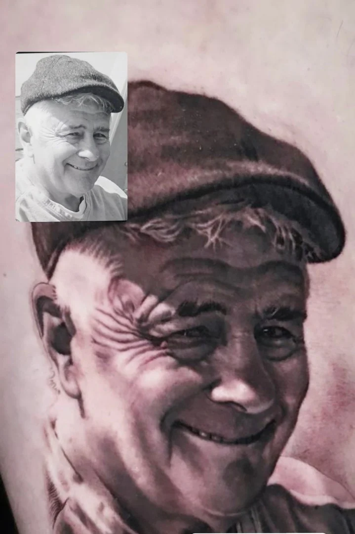 Black and grey portrait realism forearm tattoo of a smiling elderly man in a flat cap beside the reference photo at Bloodline Tattoo Phuket.