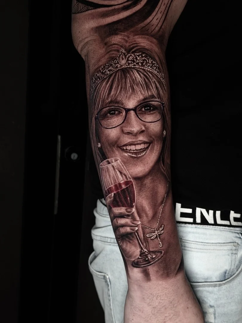Black and grey portrait realism forearm tattoo of a smiling woman with tiara, glasses, wine glass, and dragonfly necklace at Bloodline Tattoo Phuket.