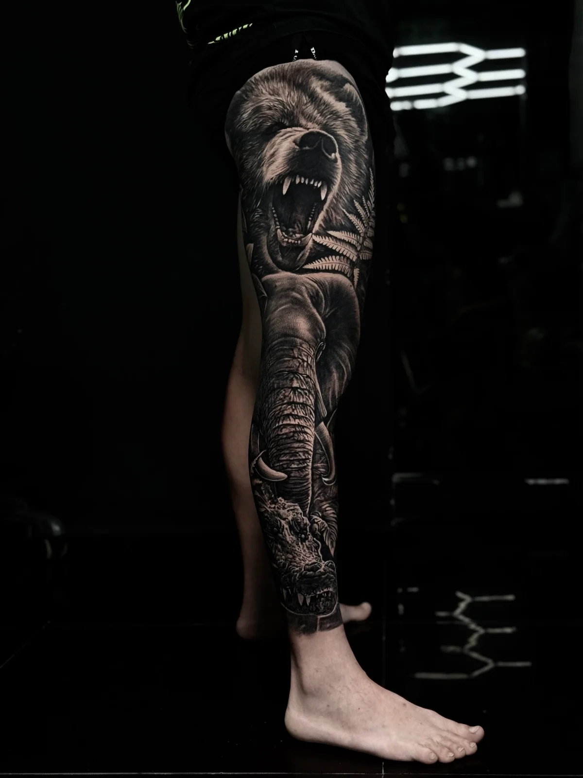 Detailed black and grey realism full leg sleeve tattoo featuring a roaring bear, an elephant, and a crocodile at Bloodline Tattoo Phuket.