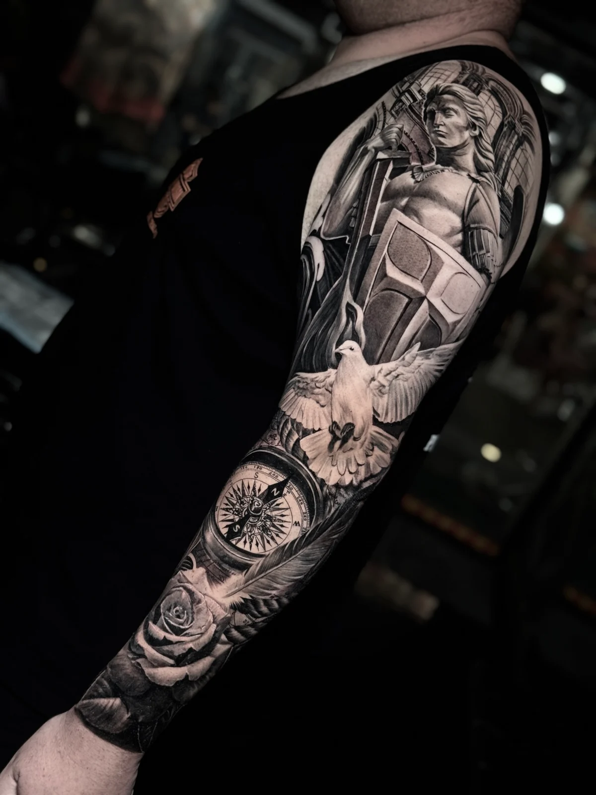 Detailed black and grey realism full sleeve tattoo featuring a classical warrior with cross shield, flying dove, compass rose, rose, and feather at Bloodline Tattoo Phuket.
