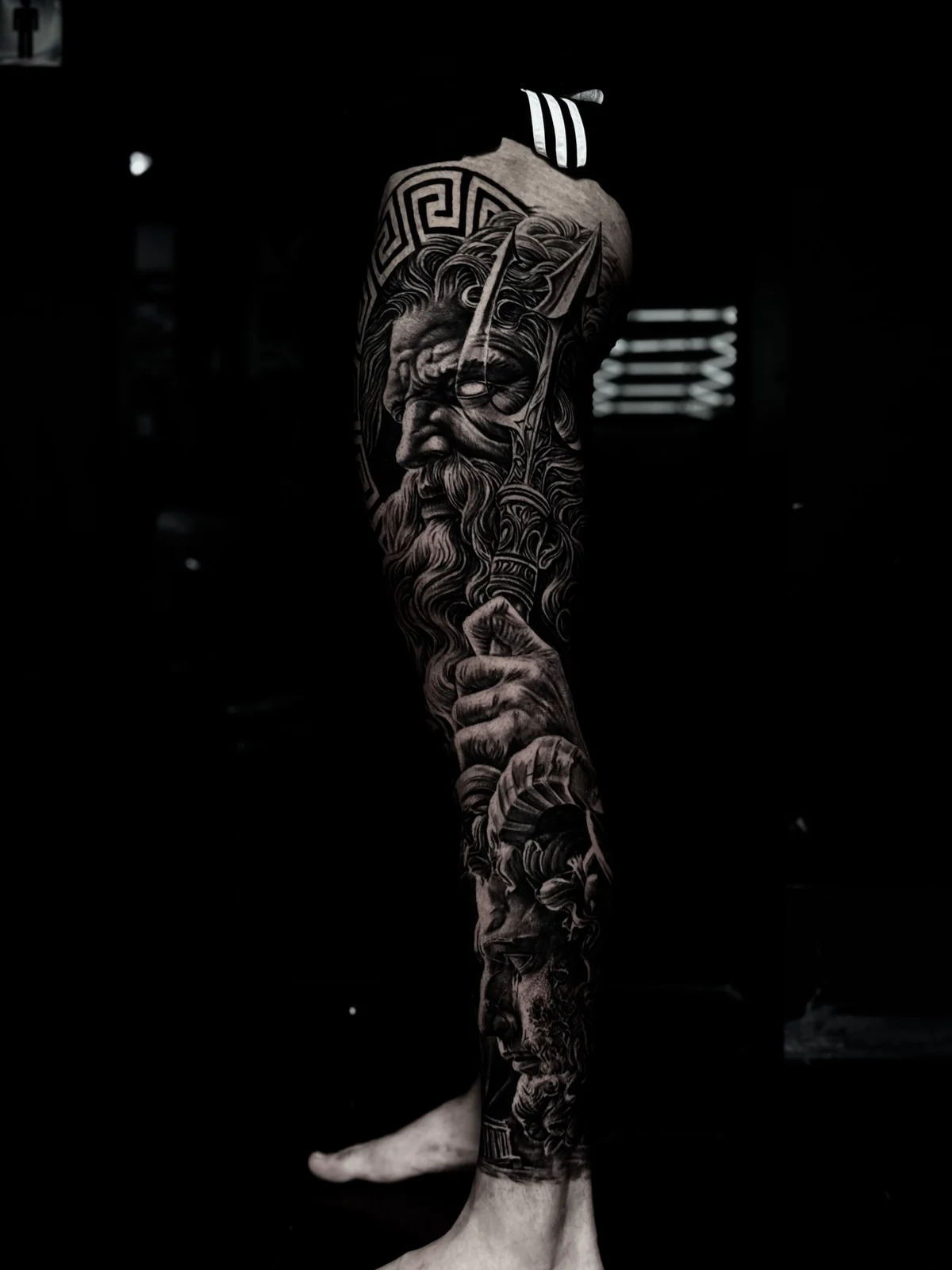 Detailed black and grey realism full leg sleeve tattoo featuring Poseidon with trident, Greek key pattern, and classical temple near the ankle at Bloodline Tattoo Phuket.